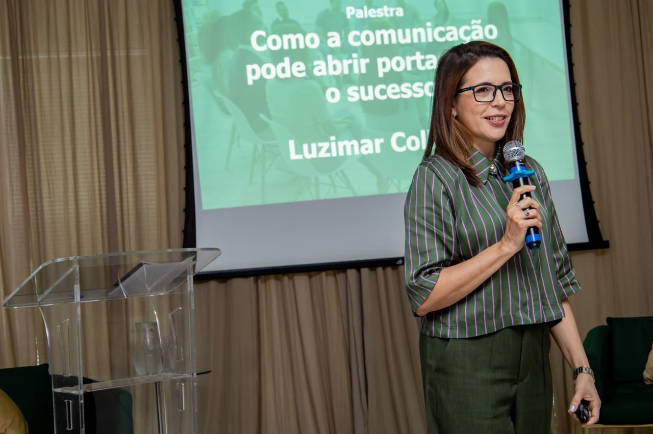 Luzimar Collares ao microfone em palestra corporativa, ao lado do telão com o tema “Como a comunicação pode abrir portas”