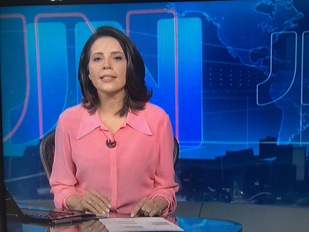 Luzimar Collares na bancada do Jornal Nacional, de blusa rosa, com o logotipo JN ao fundo